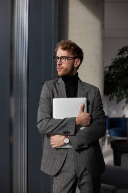 Confident young Caucasian man entrepreneur in stylish eyeglasses and formal suit, with laptop in his crossed arms, looking away in the corporate office interior. Successful people and business concept