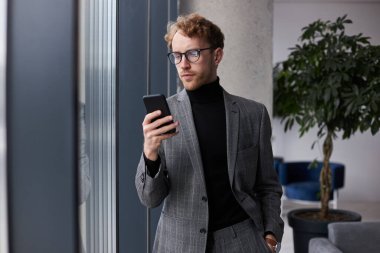 Young businessman in formal suit and eyeglasses, using his smartphone, trying new mobile applications, distant chatting, planning meeting with colleagues and business partners in the office interior