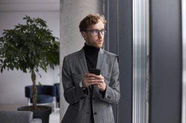 Caucasian young businessman, entrepreneur looking away while using phone, distant chatting, planning meeting with colleagues and business partners in the office interior. People Business Technology