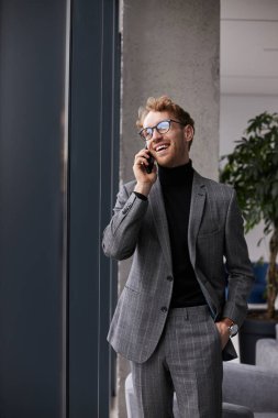Handsome confident successful male entrepreneur, developer negotiating the new actions of business project with investors on the phone, standing with his hand in the pocket in modern office interior