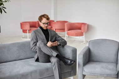 Young businessman browsing internet on his modern smartphone, sitting in the sofa in office interior. Confident developer trying new mobile application on the phone. People Technology Business concept