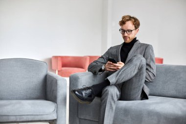 Young Caucasian man, entrepreneur freelancer, developer, wearing a gray formal business suit and eyeglasses, using mobile phone in a cozy office interior. Mobile app, technology, business concept