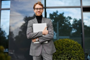 Handsome successful businessman, entrepreneur, office worker, wearing a gray business suit and stylish eyeglasses, holding laptop and smiling at camera, standing outside the corporate office building