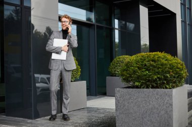 Handsome successful businessman entrepreneur in gray formal suit, smiling while negotiating plans and project with business partners on the phone, holding laptop, outside the corporate office building