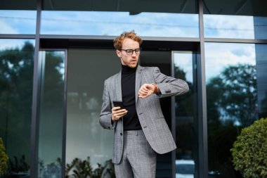 Multitasking Caucasian freelance entrepreneur, businessman holding mobile phone and checking time on wrist watch, rushing to important deal or business meeting, standing near corporate office building
