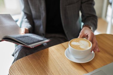 Selective focus on a male entrepreneur designer in formal wear, leafing through a catalogue while sitting at a wooden table by a cup of aroma coffee cappuccino or latte with frothed milk. Close-up.