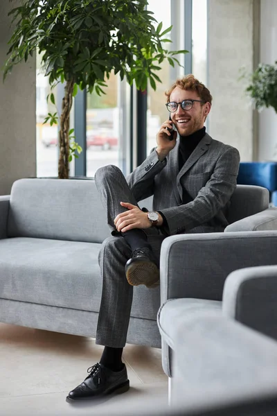 Confident businessman in gray formal suit and eyeglasses, smiles while talks on mobile phone, sitting on a comfortable sofa in the VIP lounge of a modern office interior. People Business Communication