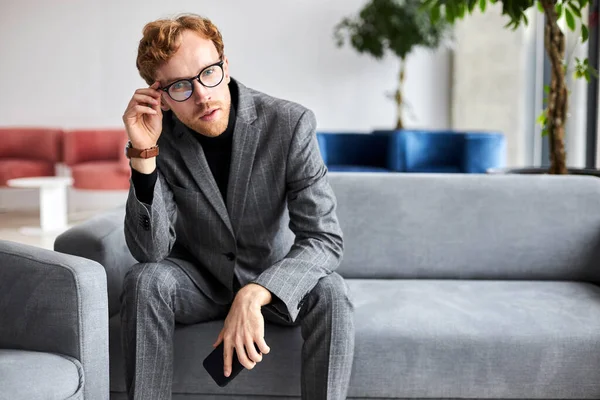 Confident multitasking Caucasian male entrepreneur, businessman, wearing eyeglasses and gray formal suit, holding smartphone in hands and looking at camera, sitting in an armchair at office reception