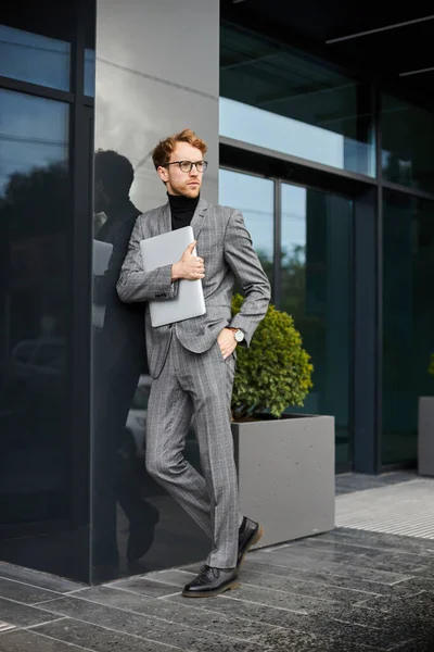 Full-length portrait of a handsome elegant male entrepreneur, businessman, wearing stylish gray business suit and eyeglasses, holding laptop in hands, looking away while outside the office building