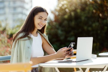 Beautiful woman holding smartphone messaging in social media sitting  in street cafe. Portrait of happy freelancer using laptop working online at workplace 