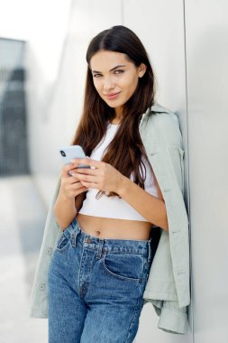 Smiling woman holding smartphone and looking at camera while standing on the street. Happy attractive female communication, chatting, creating text message. Social media concept
