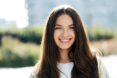 Portrait of young cheerful woman with healthy brunette hair smiling and looking at camera on street background. Happy fashion model posing outdoors. Natural beauty concept 