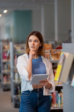 Elinde dijital tablet olan genç düşünceli bir kadın kitapçıda dikiliyor.