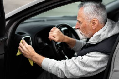 Yakışıklı, gri sakallı bir adamın yakın plan portresi. Arabasının içini sarı bir mikrofiber bezle cilalıyor ve cilalıyor. Ayrıntılar ve vale konsepti. Araba bakımı