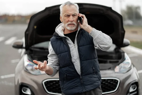 Distressed anxious senior man driver using mobile phone, calling roadside assistance, standing against the background of his car with open hood while breaking down or accident. Car insurance concept