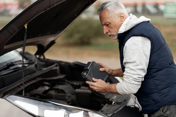 Caucasian perplexed, experienced driver, senior man adding the environmental friendly lubricant motor oil into a car engine reservoir before the ride, outdoors