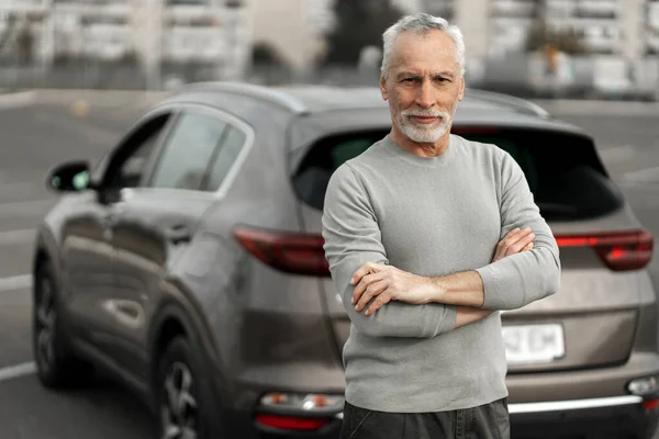 Happy senior man driver in casual clothes, smiles looking at camera, stands with his arms crossed, against the background of his newly bought car in parking lot. Lifestyle, driving and people concept