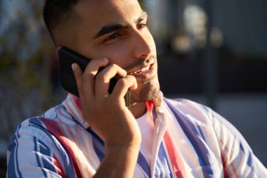 Closeup portrait of young smiling middle eastern man talking on mobile, having conversation standing on the street, selective focus. Technology concept  