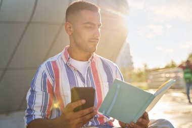 Pensive Middle eastern man holding smartphone and looking at the notebook sitting on the street. Student studying, learning something, education concept 