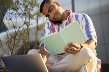 Smiling handsome middle eastern man talking on mobile phone, using laptop computer, working project sitting at workplace. Multitasking concept. Busy Iranian student studying, exam preparation 