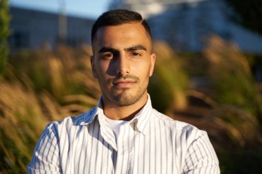 Yakışıklı, orta yaşlı, sıradan giyinmiş bir adam parkta kameraya bakıyor.