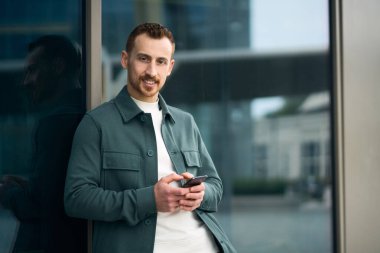 Young handsome smiling man holding smartphone using mobile app shopping online standing on the street, copy space. Hipster guy chatting outdoors, social media concept