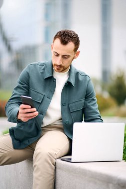 Young attractive business man checking smartphone message while working laptop at street. Online work concept
