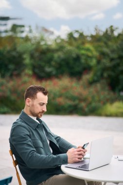Young attractive business man checking email using smartphone while working laptop online at outdoors workplace. Technology and people concept