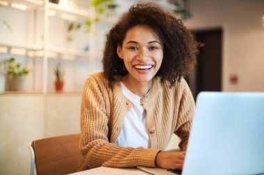 Successful young African American pretty woman, entrepreneur in casual wear, smiling with beautiful toothy smile looking at camera, telecommuting on laptop in a modern office interior. People concept.