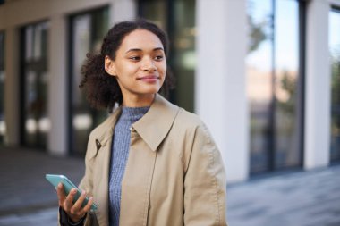 African American confident successful young woman, wearing a beige casual coat, smiles looking away, using mobile phone while walking along the street. People Technology Communication concept