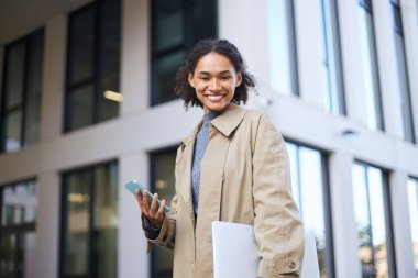 Confident successful attractive young African American businesswoman, office worker in casual coat, smiles at camera, stands with laptop and mobile phone in hands against modern urban office building