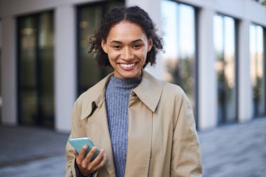 Close-up portrait of young attractive and confident multi-ethnic woman, wearing a beige casual coat, standing with a phone against a modern office building background, smiling a toothy smile at camera