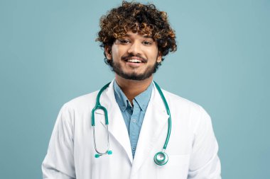 Handsome smiling Indian doctor with stethoscope wearing white lab coat isolated on blue background in hospital. Portrait of confident medical student looking at camera. Medicine, health care concept