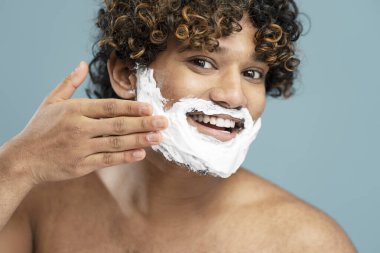 Closeup portrait of smiling bearded Indian man applying shaving foam on his face, looking at mirror in bathroom preparing for shaving. Beauty, skin care, morning routine concept 
