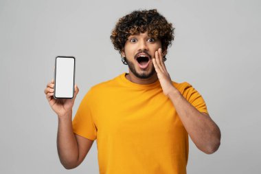 Excited Indian man with open mouth holding mobile phone, shopping online showing blank display isolated on gray background. Modern amazed asian hipster using mobile app, ordering food. Sale concept