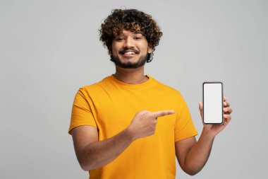 Çekici, gülümseyen, cep telefonu tutan, internetten alışveriş yapan, gri arka plandaki boş ekrana işaret eden Hintli bir adam. Modern mutlu Asyalı hipster mobil uygulama kullanıyor, yemek sipariş ediyor. Model