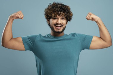 Strong smiling Indian man showing muscles, biceps looking at camera isolated on blue background. Gym workout, sport, healthy lifestyle concept 