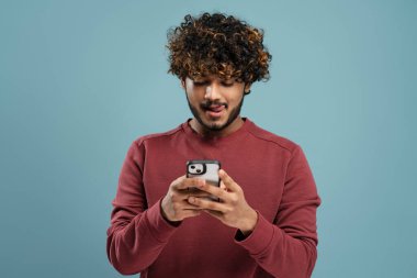 Hungry Indian man licking lips, holding smartphone using mobile app shopping online, ordering food on website isolated on grey background, copy space