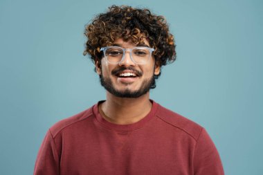 Portrait of attractive smiling curly haired Indian man wearing stylish eyeglasses looking at camera isolated on blue background. Vision concept 