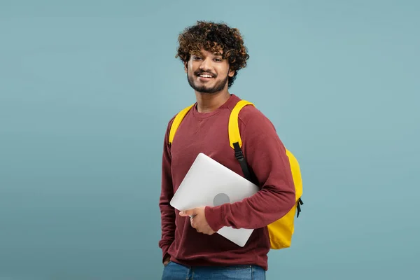 Attractive smiling Indian student with yellow backpack holding laptop, looking at camera isolated on blue background. Education concept