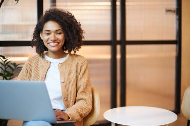 Young African American successful woman in casual wear, working remotely on laptop and smiling a beautiful toothy smile looking at camera. People, business, career and professional ambitions concept
