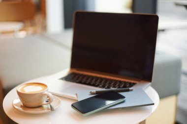Still life with mobile phone and laptop with blank black monitor screen, near a white cup of coffee latte with frothed foam, on a small table in the office interior. Business and coffee break concept