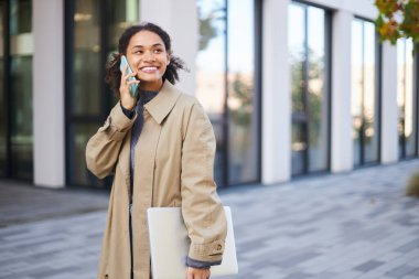 Attractive happy African American young woman in beige casual coat, smiling and talking on mobile phone while strolling the city street with laptop in her hands. People Lifestyles Business Technology