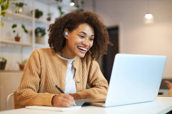 Happy young African American female confident student or university graduated, smiling, watching webinar in online class, taking notes on notebook. People. Online education. Distance learning concept