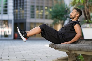 Fit man pumping press sitting on bench. Muscular man doing his morning exercises. Black sport clothes. Street on background 