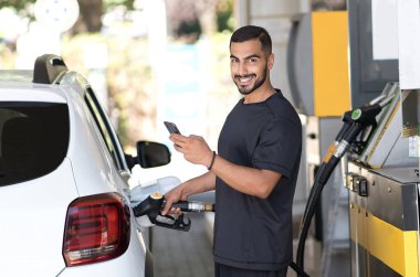 Smiling man refuelling his luxury car and messaging smartphone at the gas station. Iranian guy looking camera and pouring petrol into tank of modern vehicle on filling station in city 