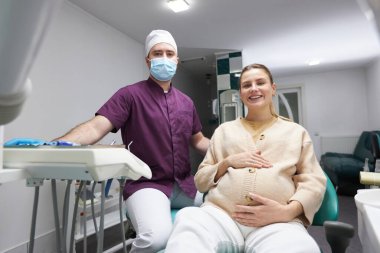 Male dentist doctor and beautiful pregnant woman smiling a cheerful toothy smile, looking at camera after regular dental check-up or teeth treatment in the dental office. People. Service. Healthcare