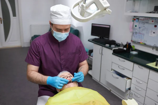 歯科医院の歯医者室の若い女性患者の口腔と歯茎を調べ 歯科用鏡とステンレス製のツールを使用して 医療用マスクと制服を着た男性歯科衛生士 — ストック写真