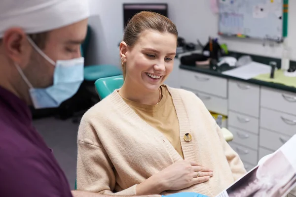 在与牙科医生预约的时候 年轻的孕妇把手放在肚子上 现代牙科诊所的X光全景图像解释说 正牙医生戴着医疗面罩 — 图库照片