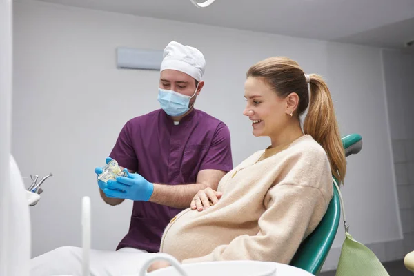 孕妇坐在牙医的椅子上 听医生向她解释人类颌骨模型时 脸上挂着可爱的笑容 这是在牙科诊所里需要做的治疗 — 图库照片